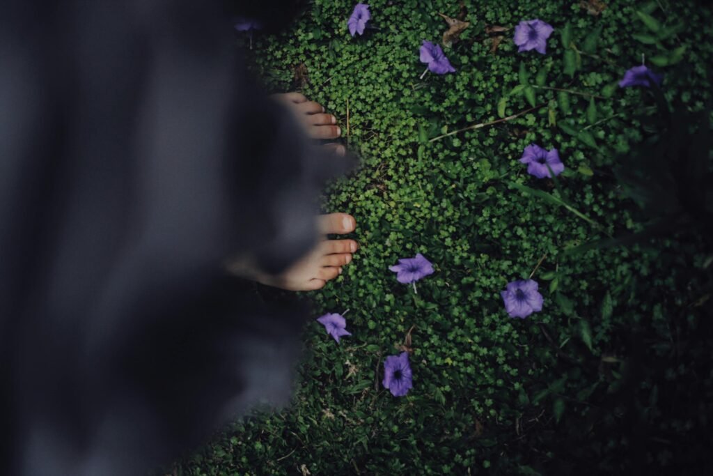 A person stands barefoot on green grass surrounded by purple flowers, creating a serene outdoor scene.